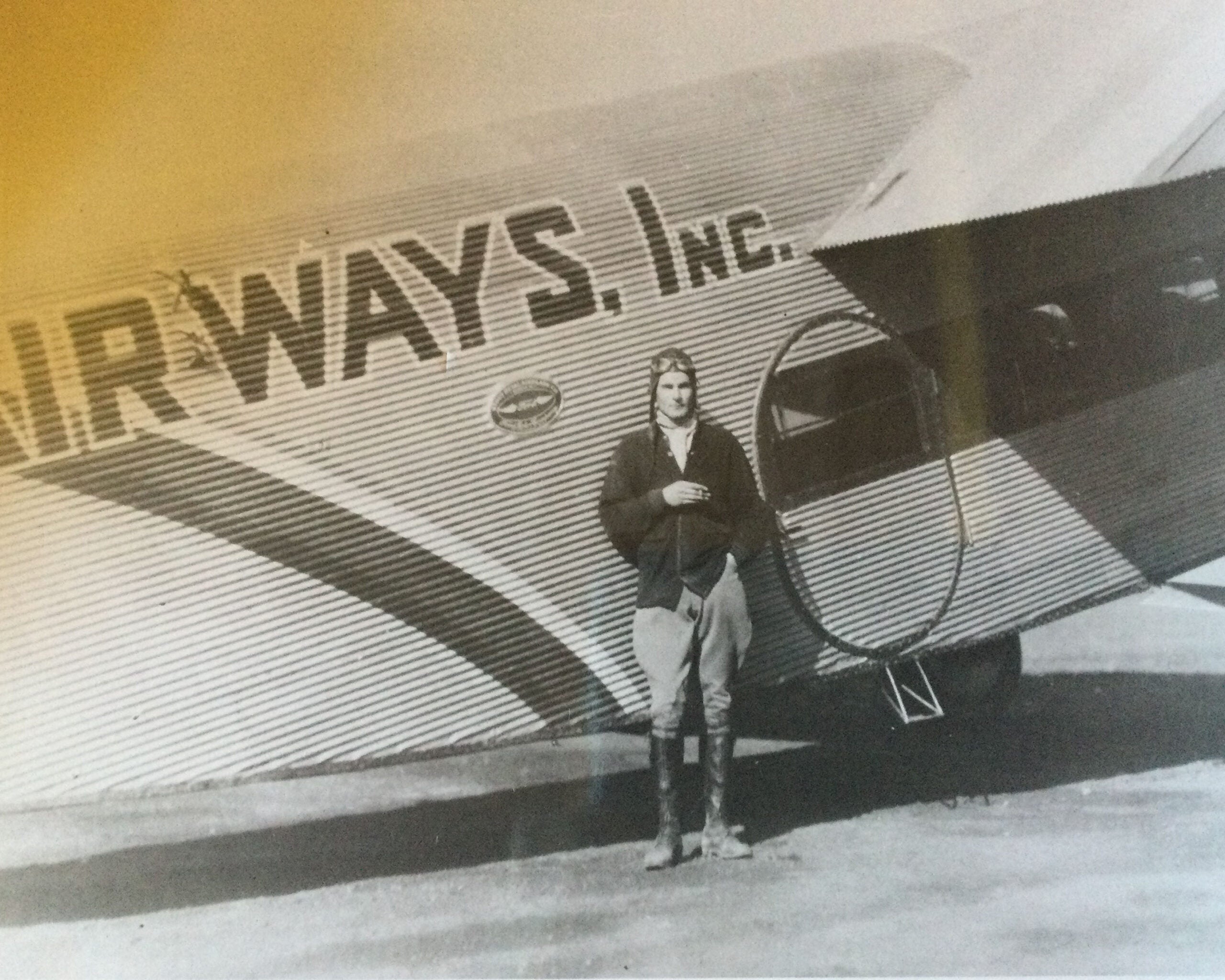 A person in aviator attire stands in front of a vintage airplane with AIRWAYS, INC. written on its side.