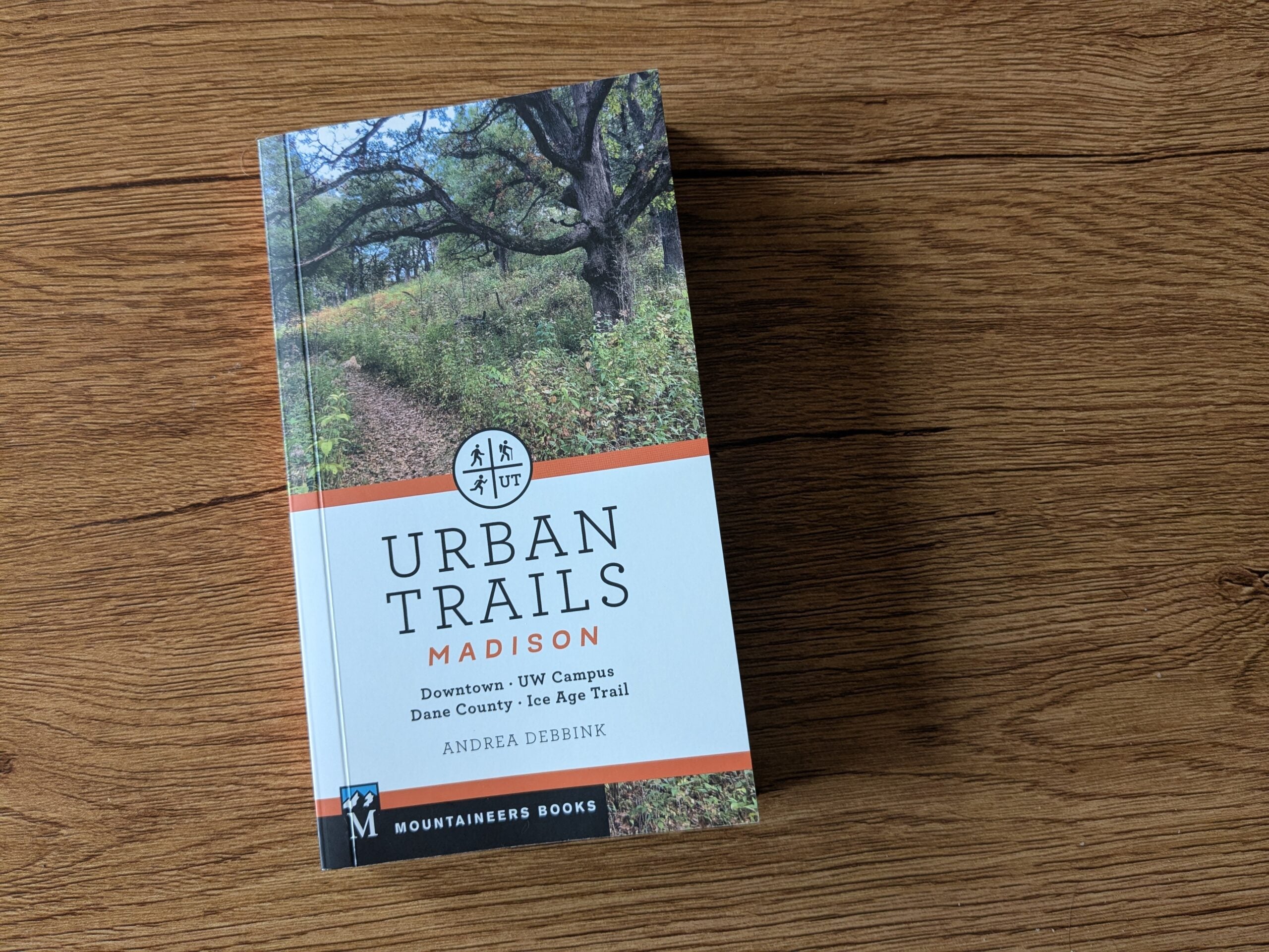 A book titled Urban Trails: Madison by Andrea Debbink rests on a wooden surface. The cover features a photo of a wooded trail.