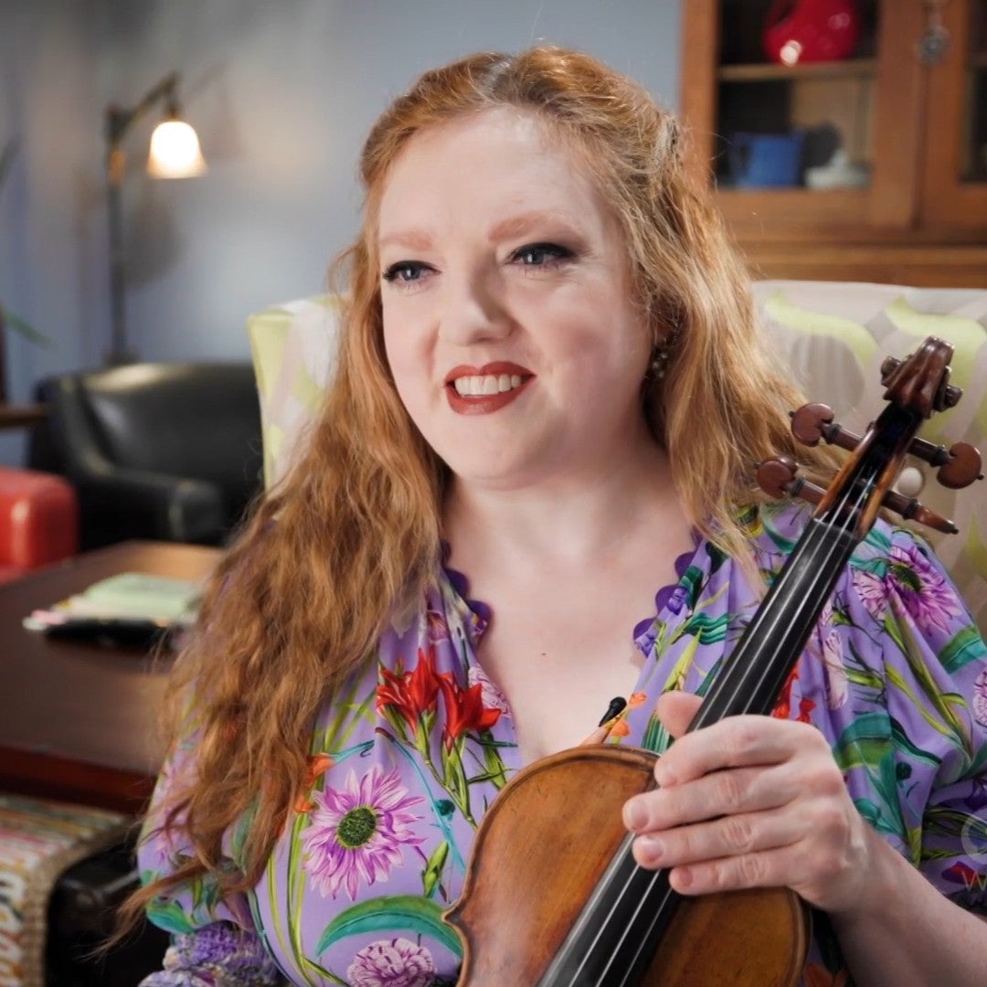 A woman with long red hair holds a violin while seated indoors, smiling at the camera. She is wearing a floral-patterned blouse.