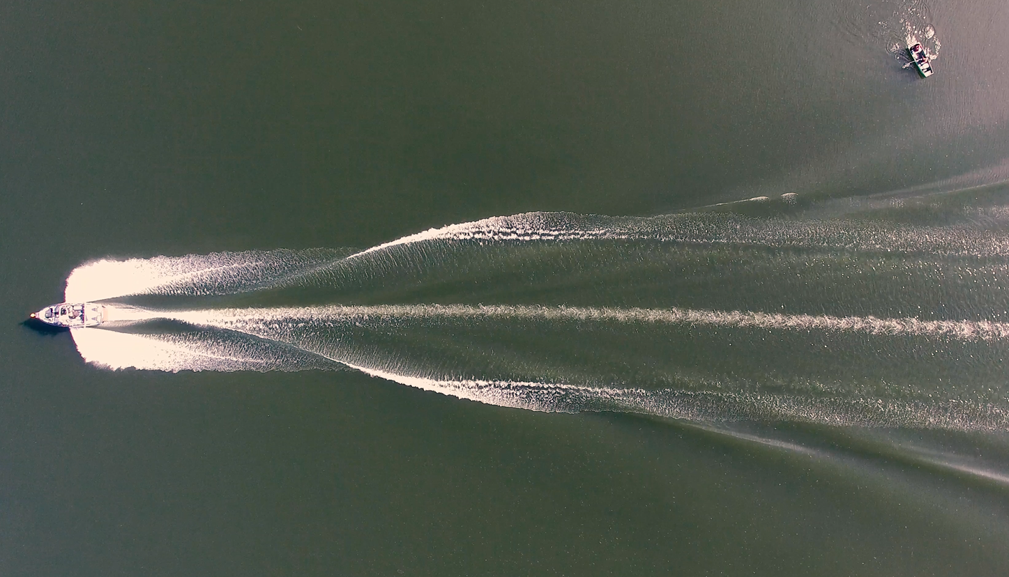 Aerial view of a speedboat creating a V-shaped wake in the water, with a smaller boat visible in the distance on the upper right.