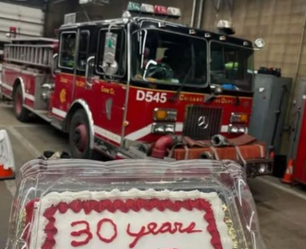 A fire truck is parked in a garage behind a cake with 30 years written in red icing on top.