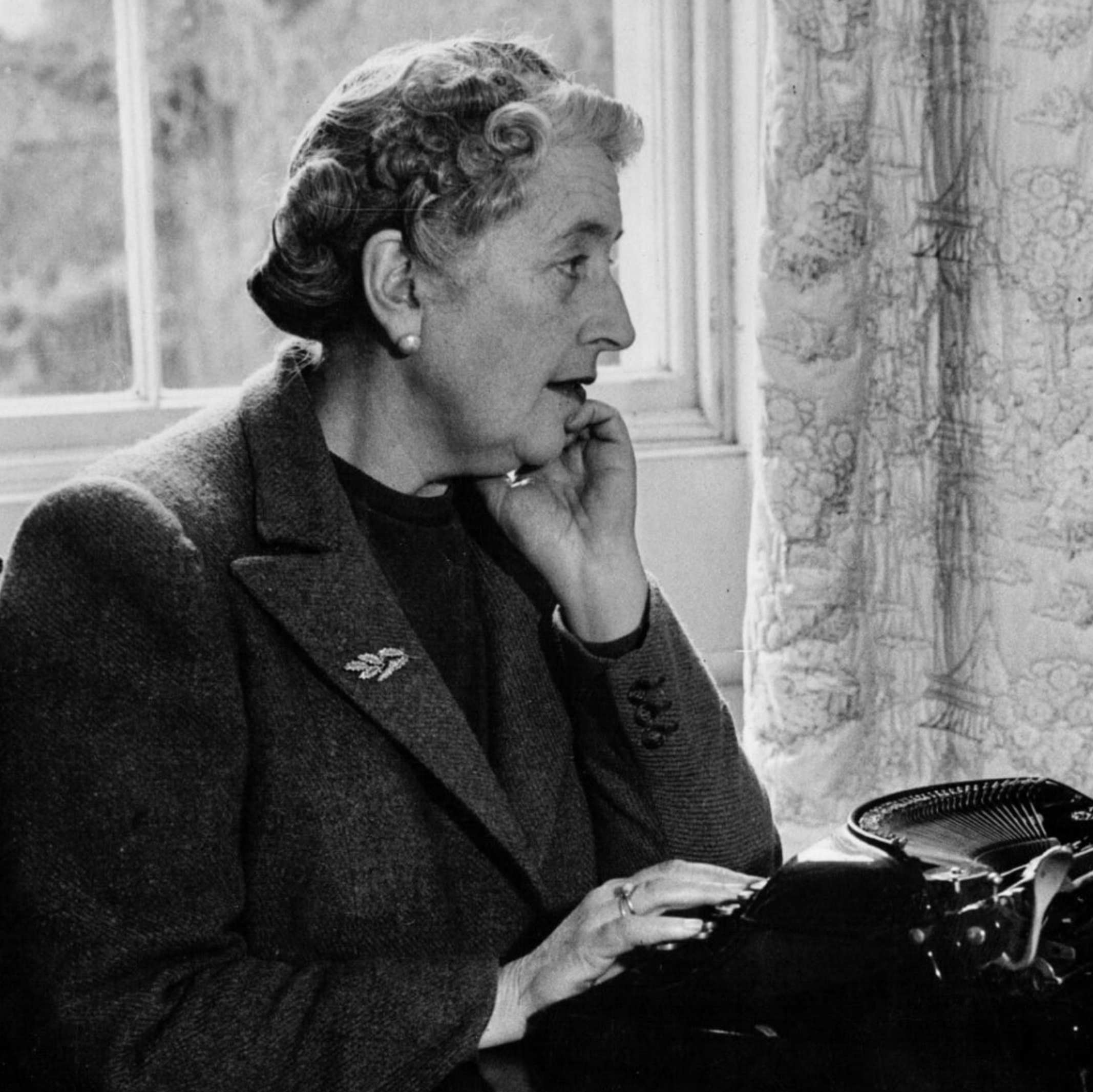 An older woman with short, curly hair sits at a desk by a window, touching her face with one hand and using a typewriter with the other. A patterned curtain and trees are visible behind her.