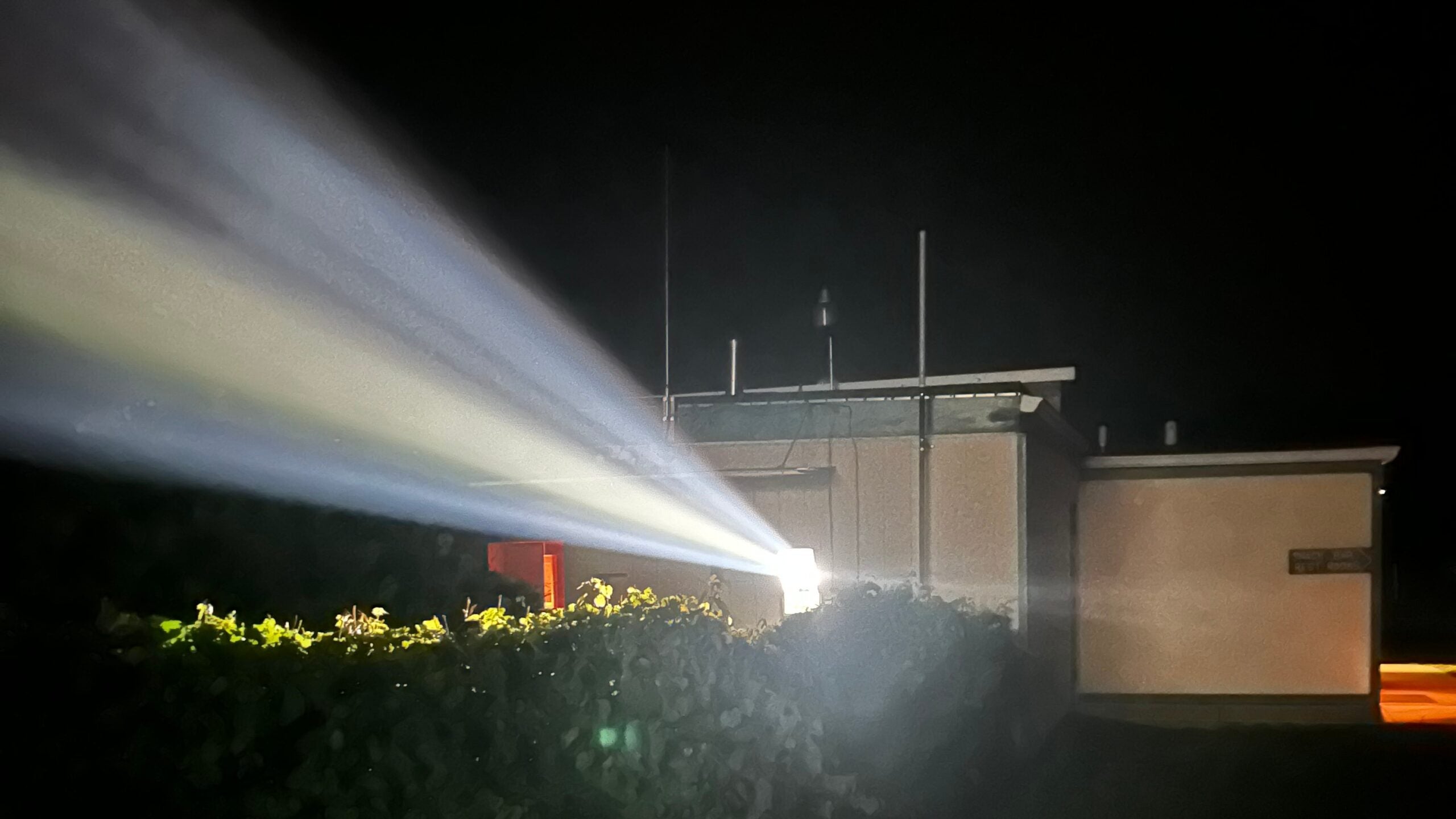 A bright light projects strong beams across foliage toward a small building at night.