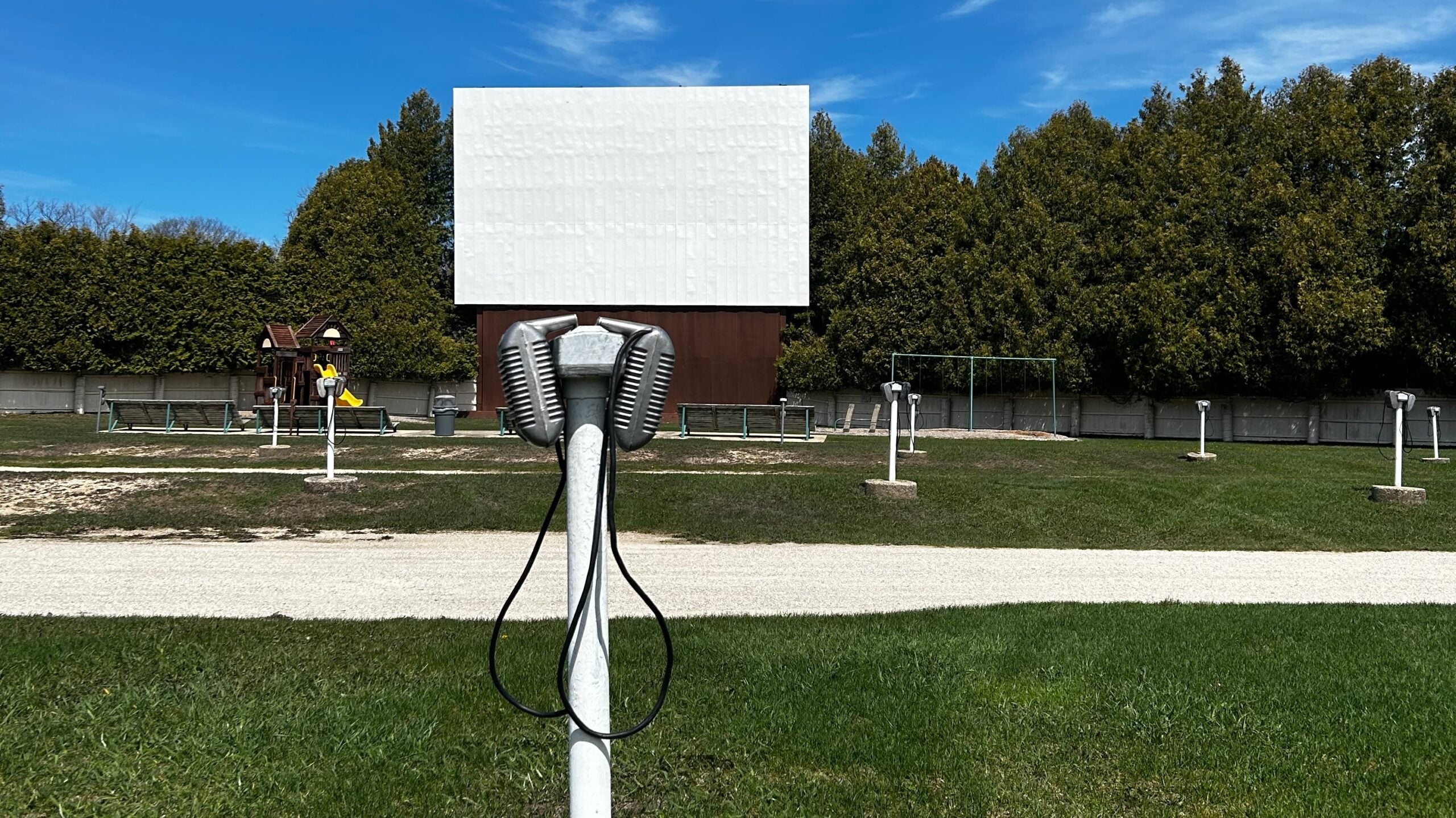 A retro outdoor drive-in movie theater with a blank white screen, classic metal speaker in the foreground, and rows of parking spots on green grass.