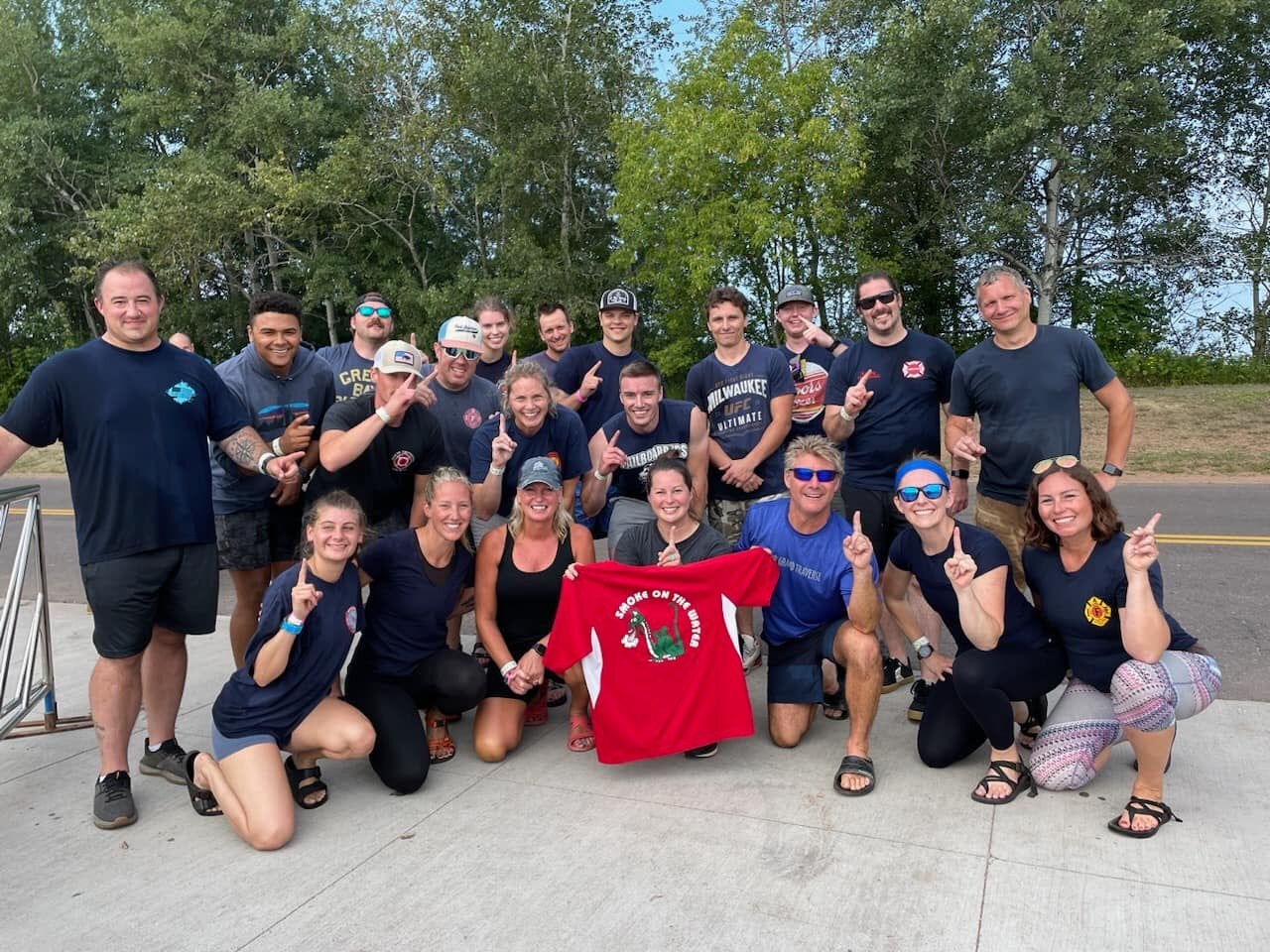 A group of people pose outdoors on a sidewalk, some kneeling and some standing, holding up one finger. One person in front holds a red shirt. Trees are visible in the background.