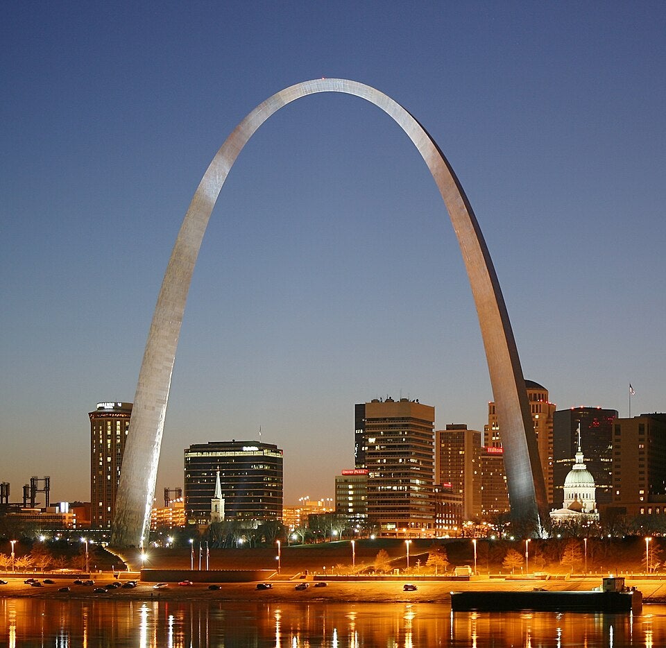 The Gateway Arch in St. Louis, Missouri, illuminated at dusk with city buildings and the Mississippi River in the foreground.