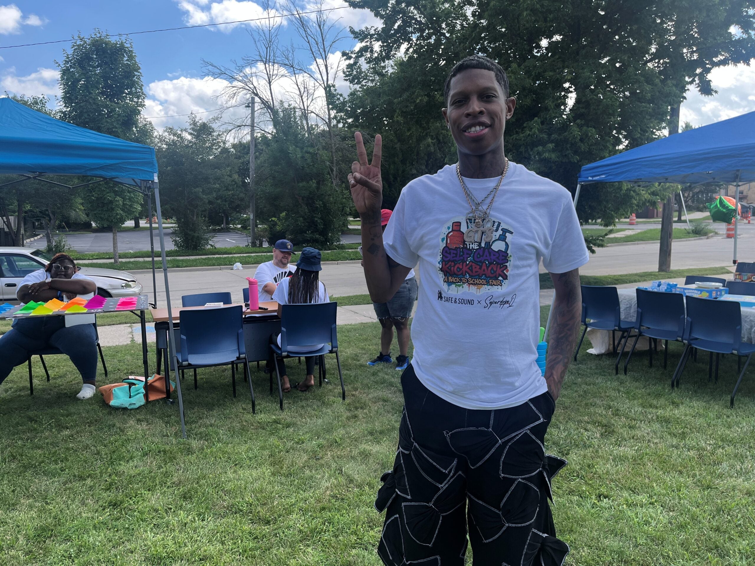A man stands on grass smiling and flashing a peace sign at an outdoor event; people sit at tables under blue canopy tents in the background.