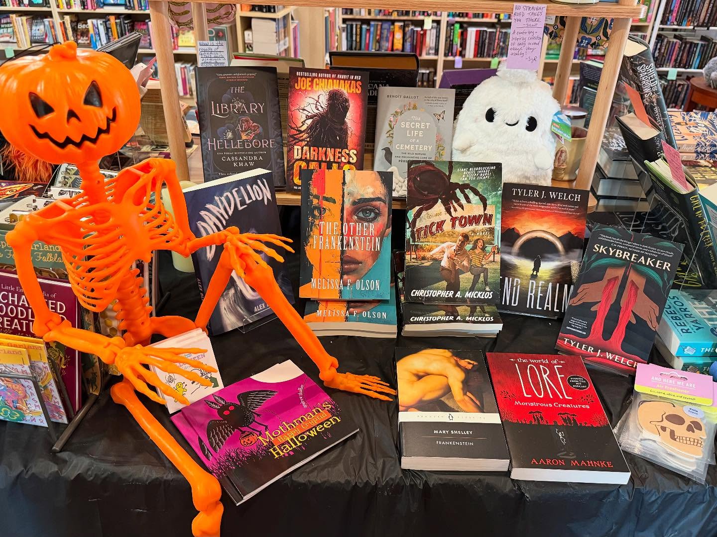 A display table in a bookstore featuring Halloween-themed books, a bright orange skeleton, a pumpkin head figure, and a white plush ghost among the shelves of books.