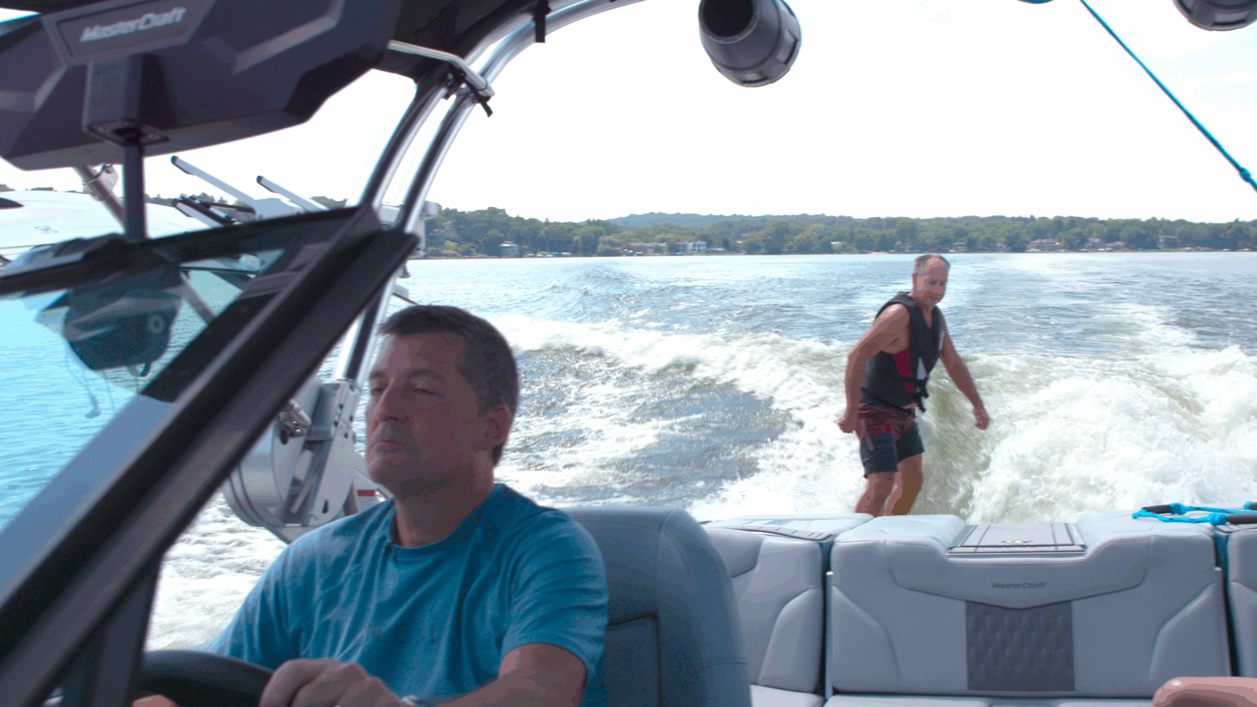 Man drives a boat while another man in a life vest wakesurfs behind, with water and shoreline visible in the background.