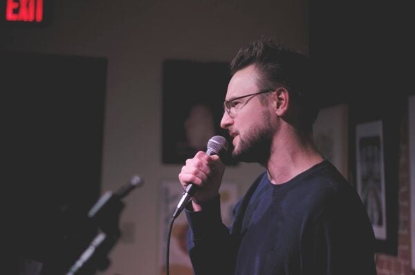 A man in glasses holds a microphone and speaks on stage, with a blurred background and an exit sign visible.