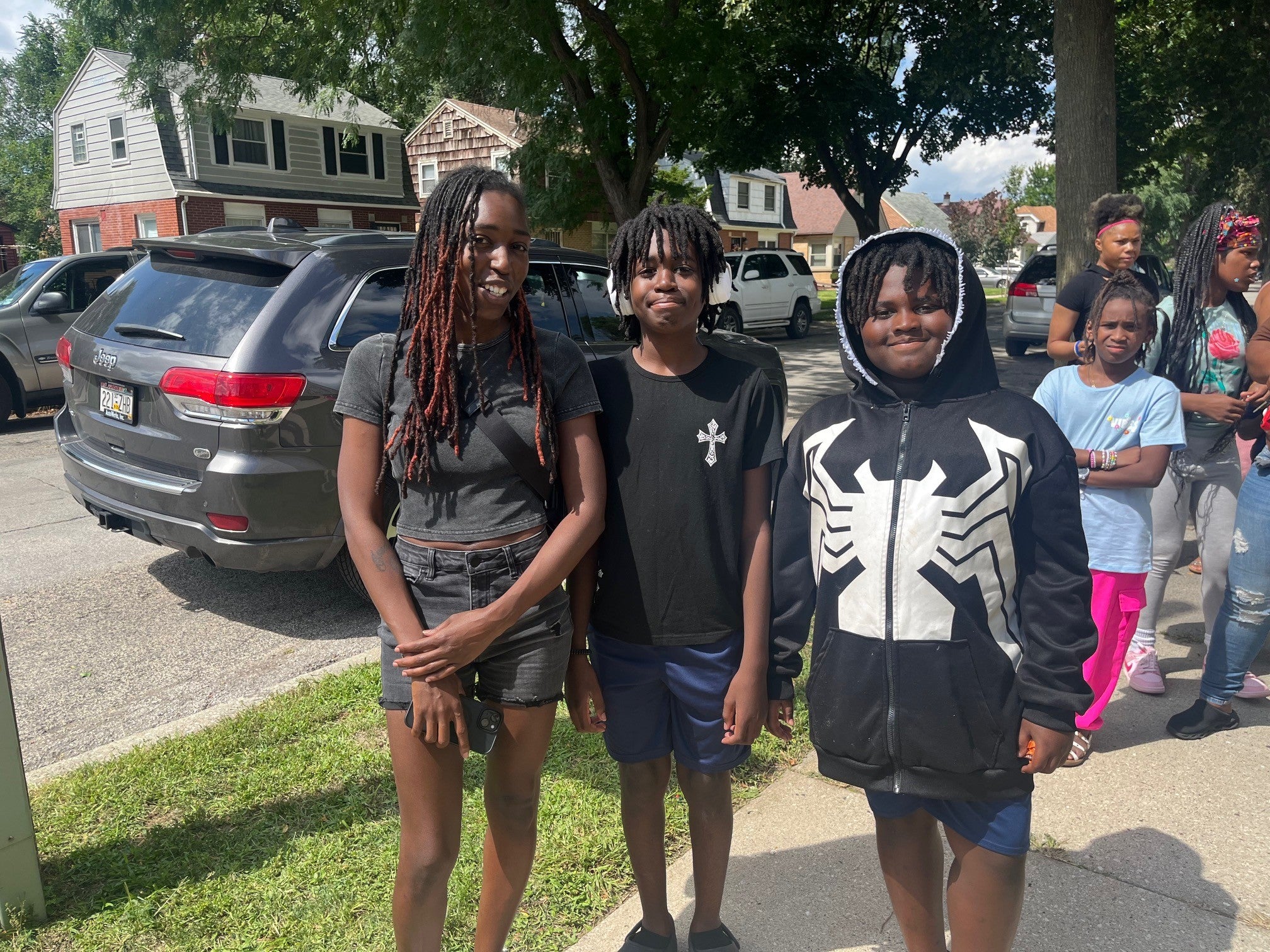 Three kids stand on a sidewalk in front of parked cars and houses on a sunny day; others are gathered in the background on the grass and pavement.