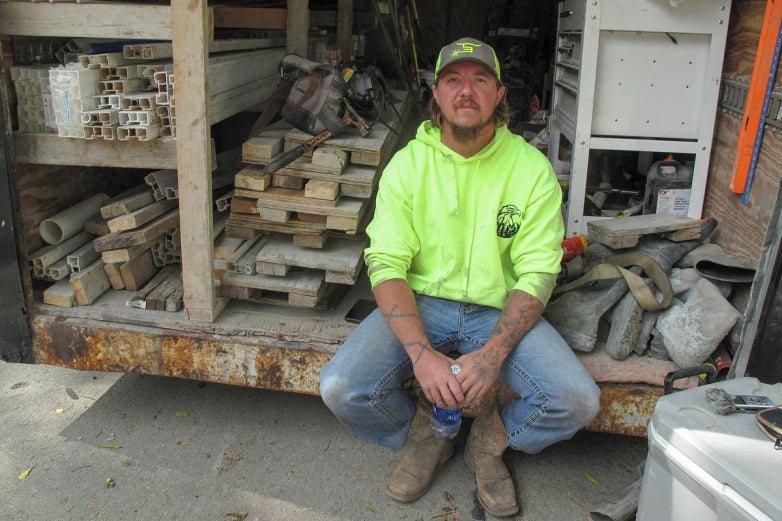 A man in a neon yellow hoodie and cap sits on the edge of a work truck loaded with various wooden planks and construction materials.