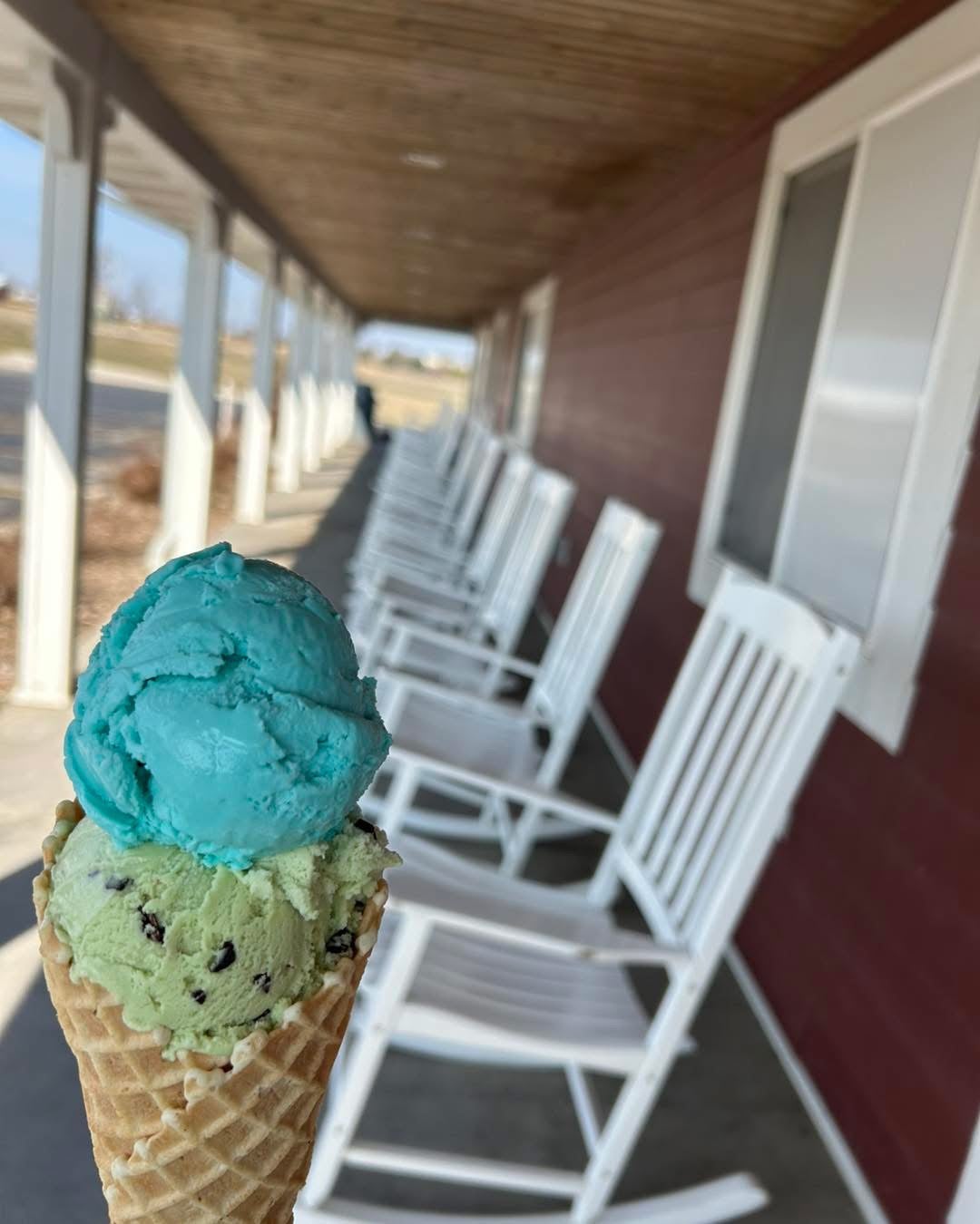 A hand holds an ice cream cone with blue and green scoops in front of a row of empty white rocking chairs on a covered porch.