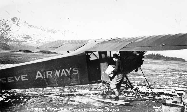 Black and white photo of a man unloading freight from a Reeve Airways airplane on a muddy airstrip in Valdez, Alaska, with mountains in the background.