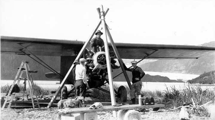 Three men work on the engine of a small airplane supported by a wooden tripod outdoors, with mountains and a glacier in the background.