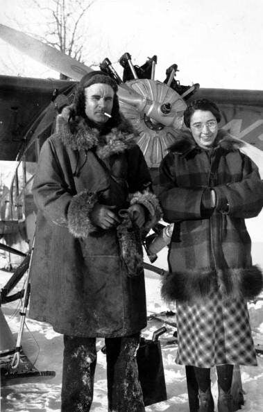A man in fur clothing and a woman in a checkered coat stand in front of a propeller plane in a snowy setting.