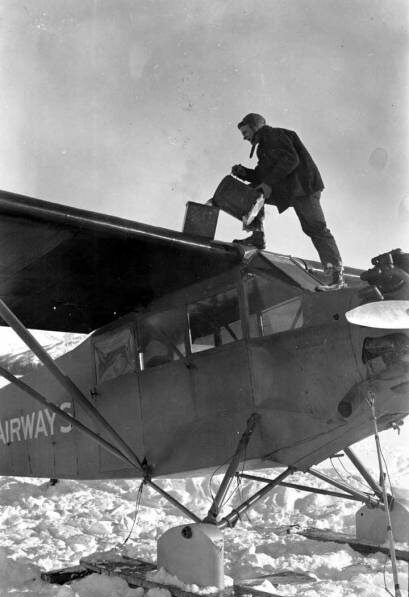 A person stands on the wing of a small airplane, pouring liquid from a can into a tank, surrounded by snow-covered ground.