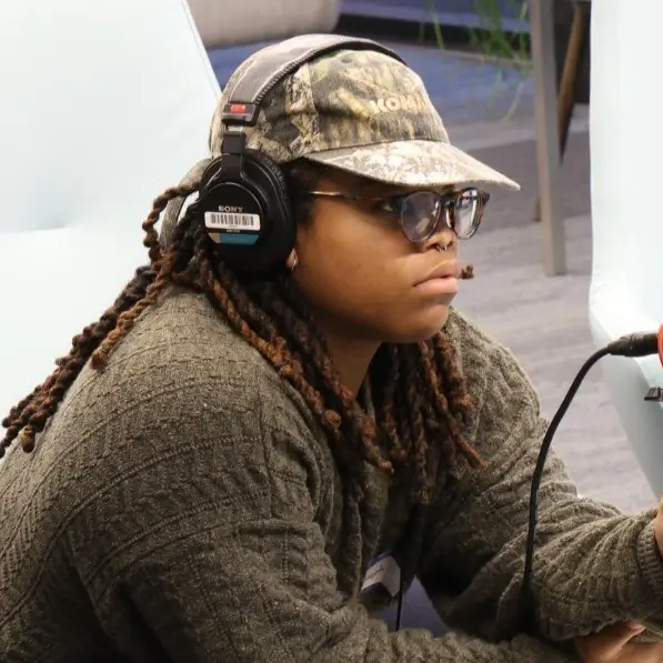 A person wearing a gray sweater, camouflage baseball cap, sunglasses, and headphones is sitting indoors, looking focused. A microphone is positioned in front of them.