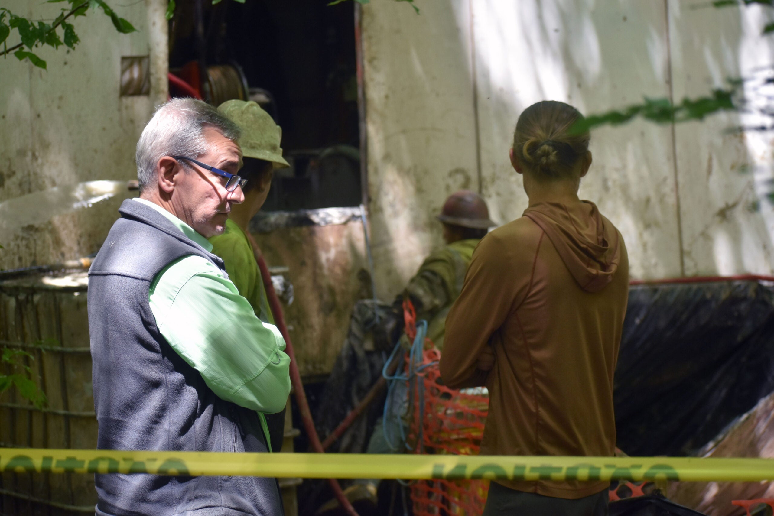 Several people stand behind caution tape at a work site, near construction materials and equipment, with trees and a white structure in the background.