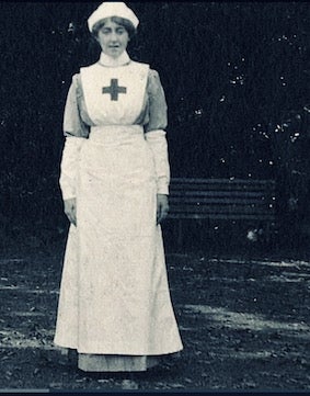 A woman wearing a traditional nurse uniform with a Red Cross emblem stands outdoors in front of a bench.
