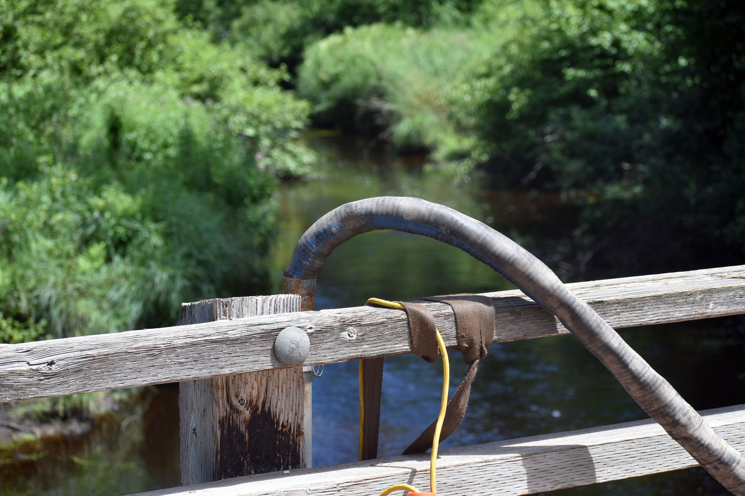 A close-up of a weathered wooden railing with a hose and rope draped over it, overlooking a creek surrounded by green foliage.