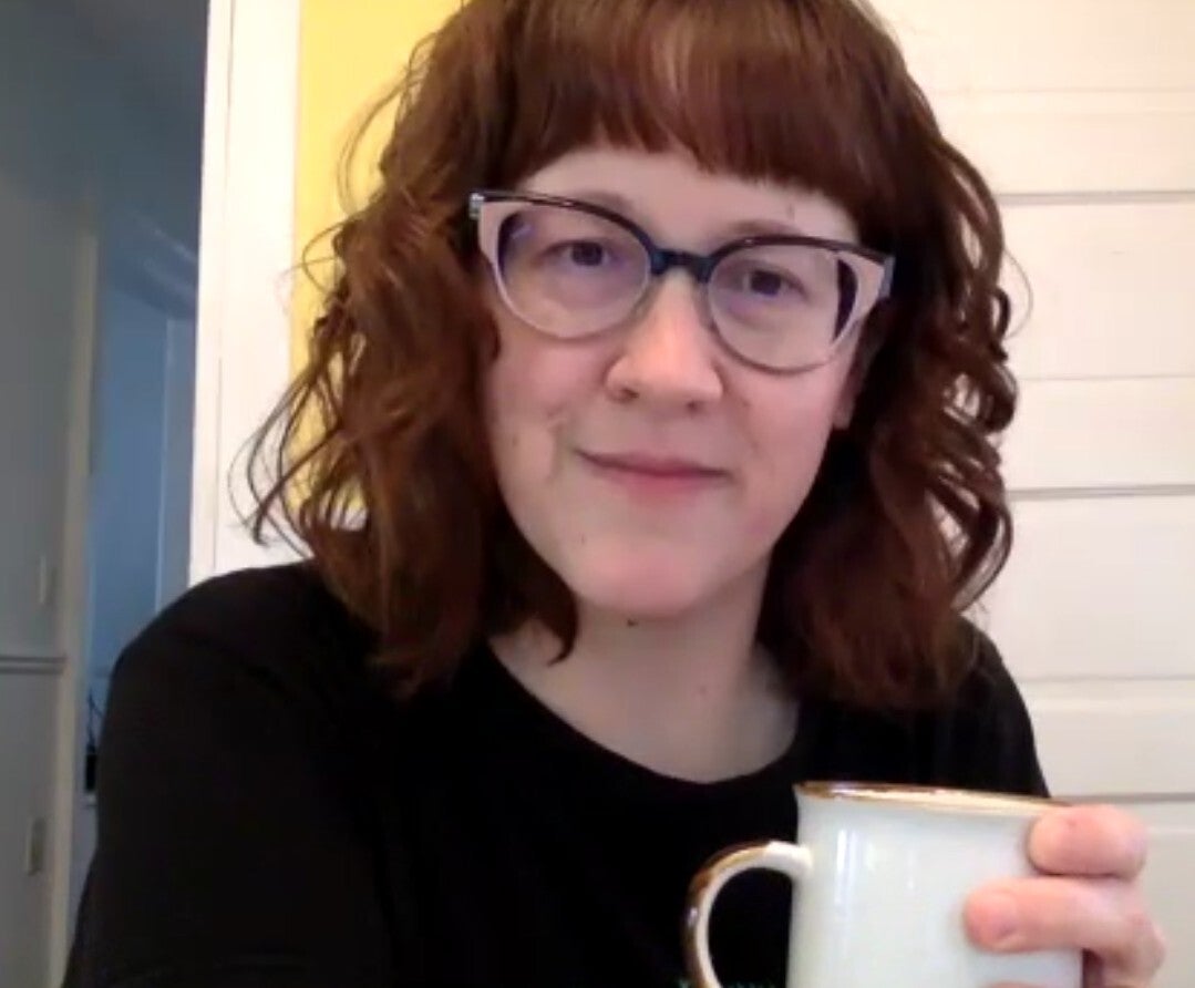 Person with wavy brown hair and glasses holding a white mug, wearing a black top, sitting indoors in front of a white door and yellow wall.