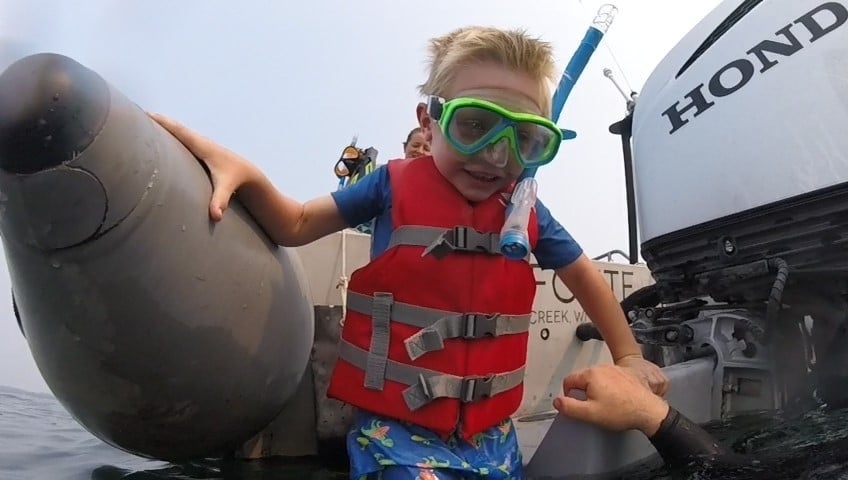 A young boy wearing a snorkel, goggles, and a red life vest climbs onto a boat from the water, assisted by an adult’s hand.