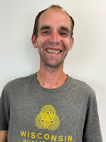 A man with short brown hair and a receding hairline smiles at the camera, wearing a gray Wisconsin T-shirt with a yellow graphic, standing before a plain white wall.