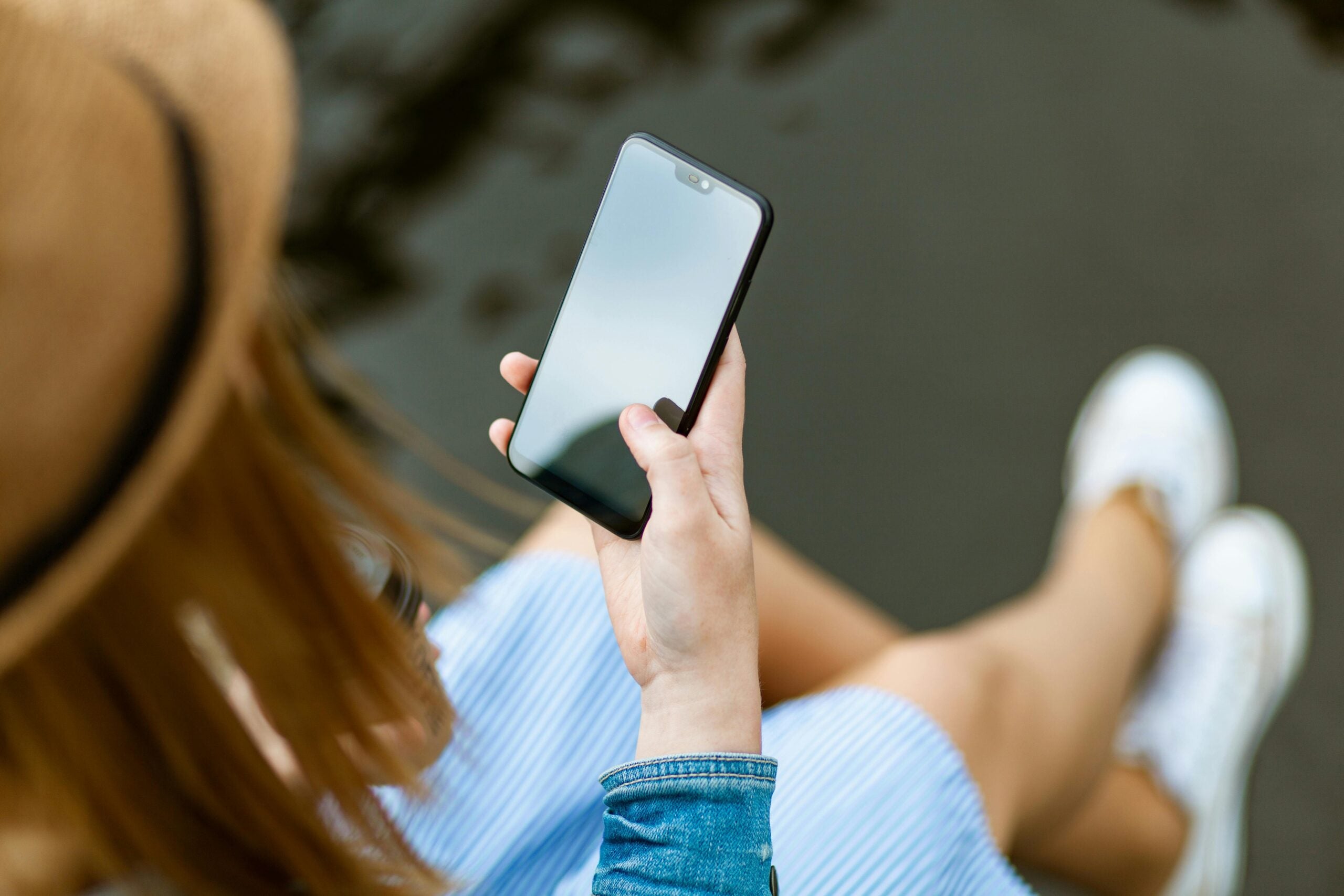A person wearing a hat and blue striped dress is sitting and holding a smartphone, looking at its blank screen.
