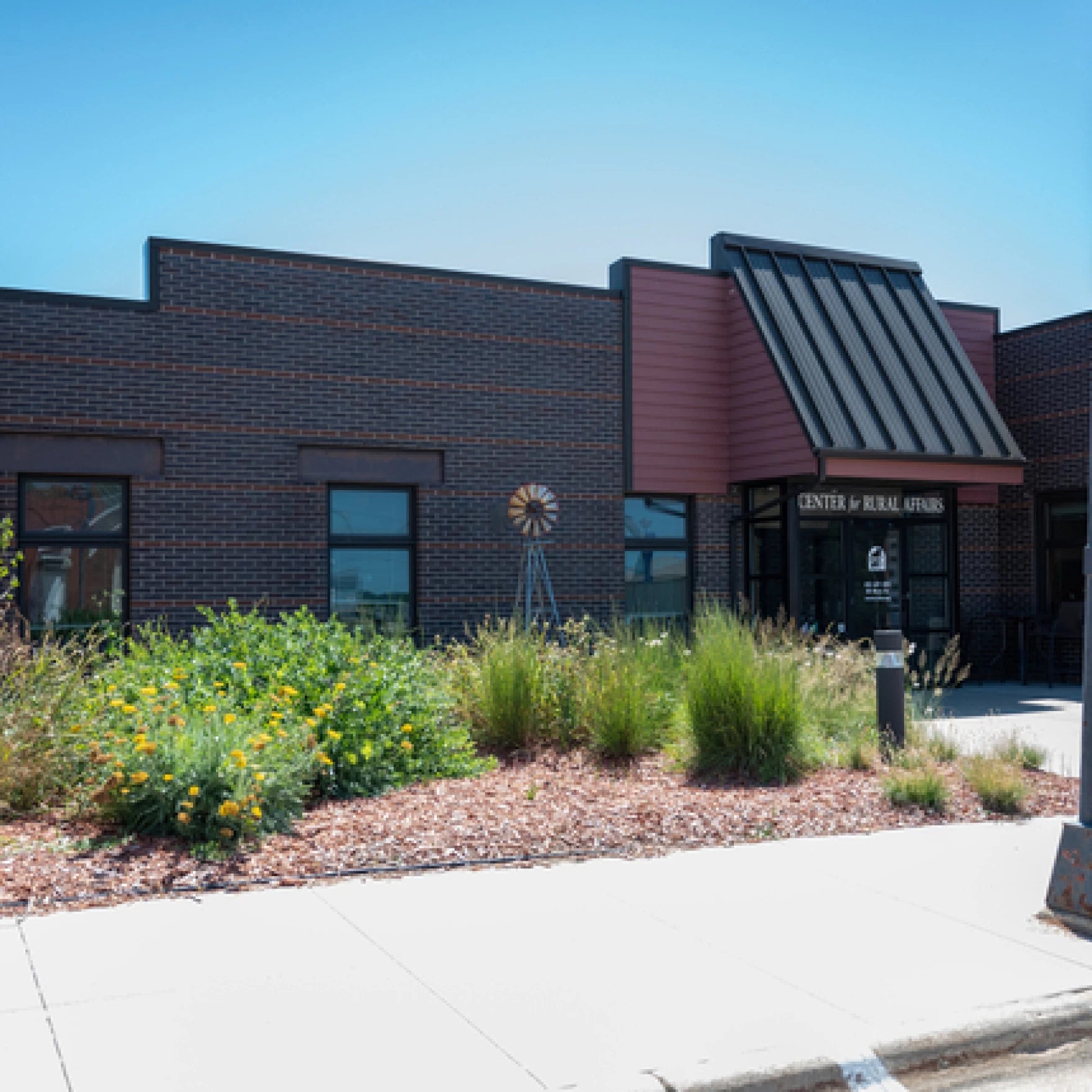 Single-story brick building with a black metal awning, labeled Center for Rural Affairs, landscaped with native plants and a sidewalk in front.