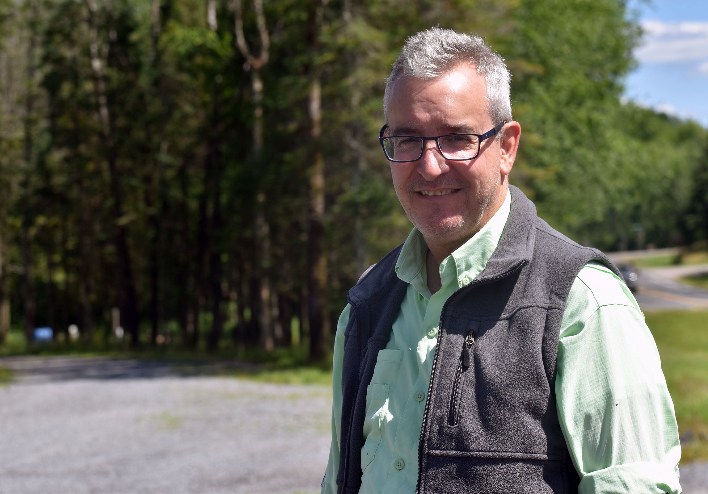 A man with gray hair and glasses stands outdoors near a road, wearing a light green shirt and dark vest, with trees and greenery in the background.