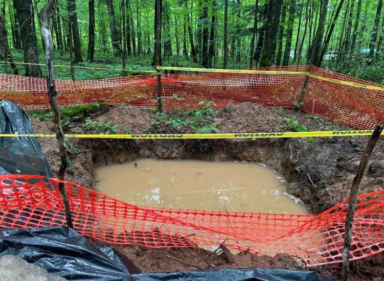 A muddy water-filled pit is surrounded by orange safety fencing and caution tape in a wooded area.