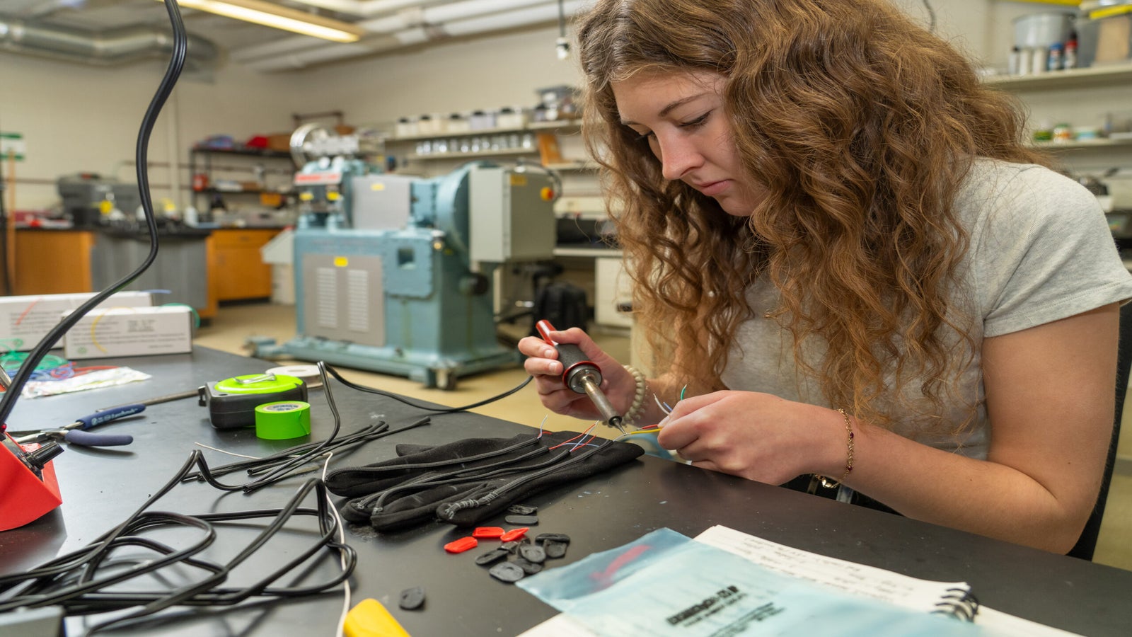 A woman uses a soldering iron to work on electronic components at a lab workstation, with tools and equipment visible in the background.