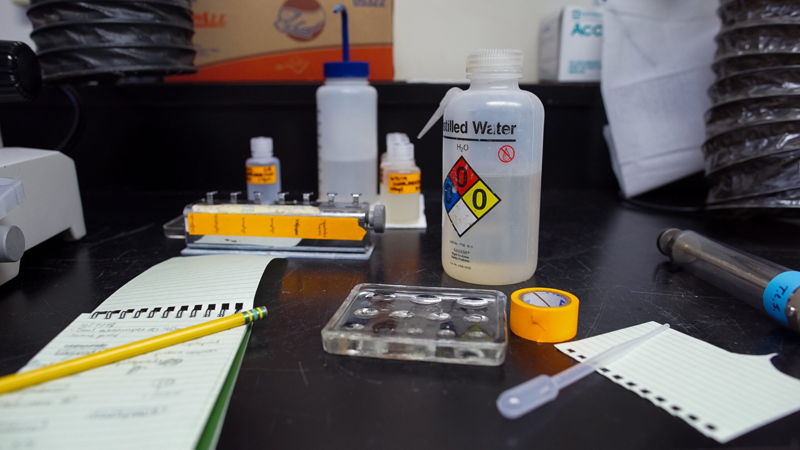 A lab bench with distilled water, pipette, tape, notepads, pencil, a small tray with wells, and labeled bottles organized for an experiment.