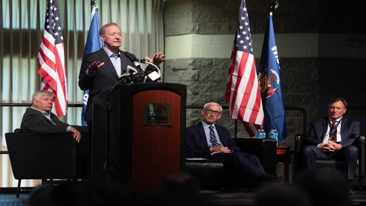 Four men in suits are on stage at a panel event; one man is speaking at a podium while the other three are seated. Multiple U.S. flags are displayed in the background.