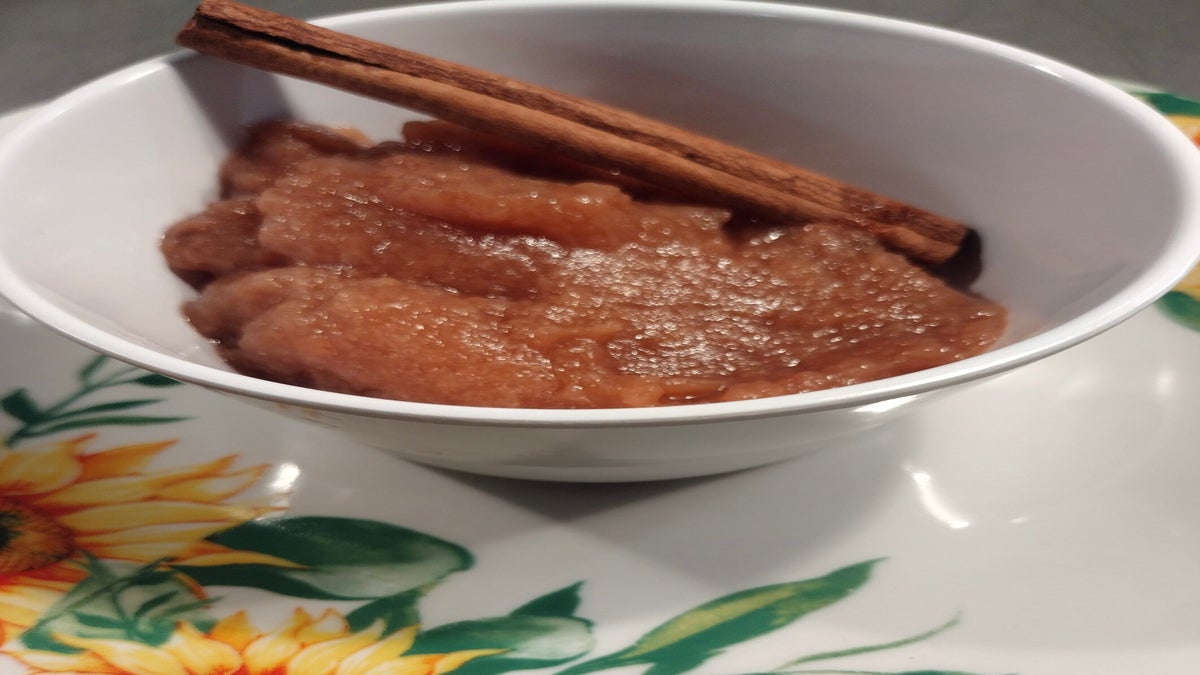 A white bowl filled with applesauce topped with a cinnamon stick, placed on a plate with a sunflower design.