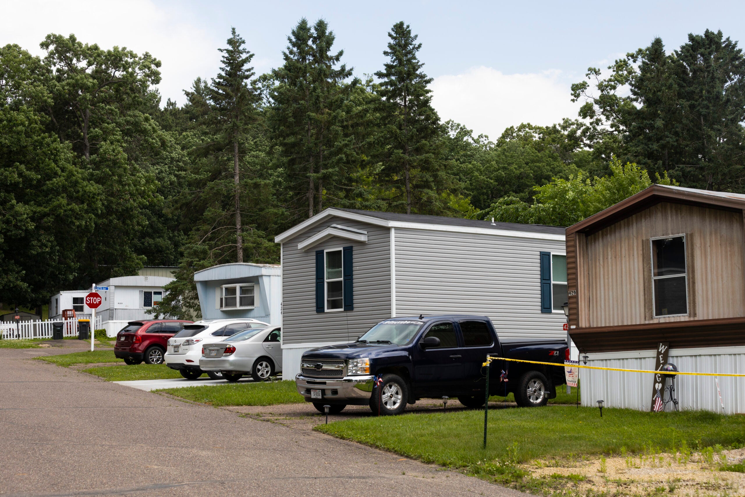 Several mobile homes are lined up along a paved road, with parked cars and trucks in front; trees and greenery surround the area.