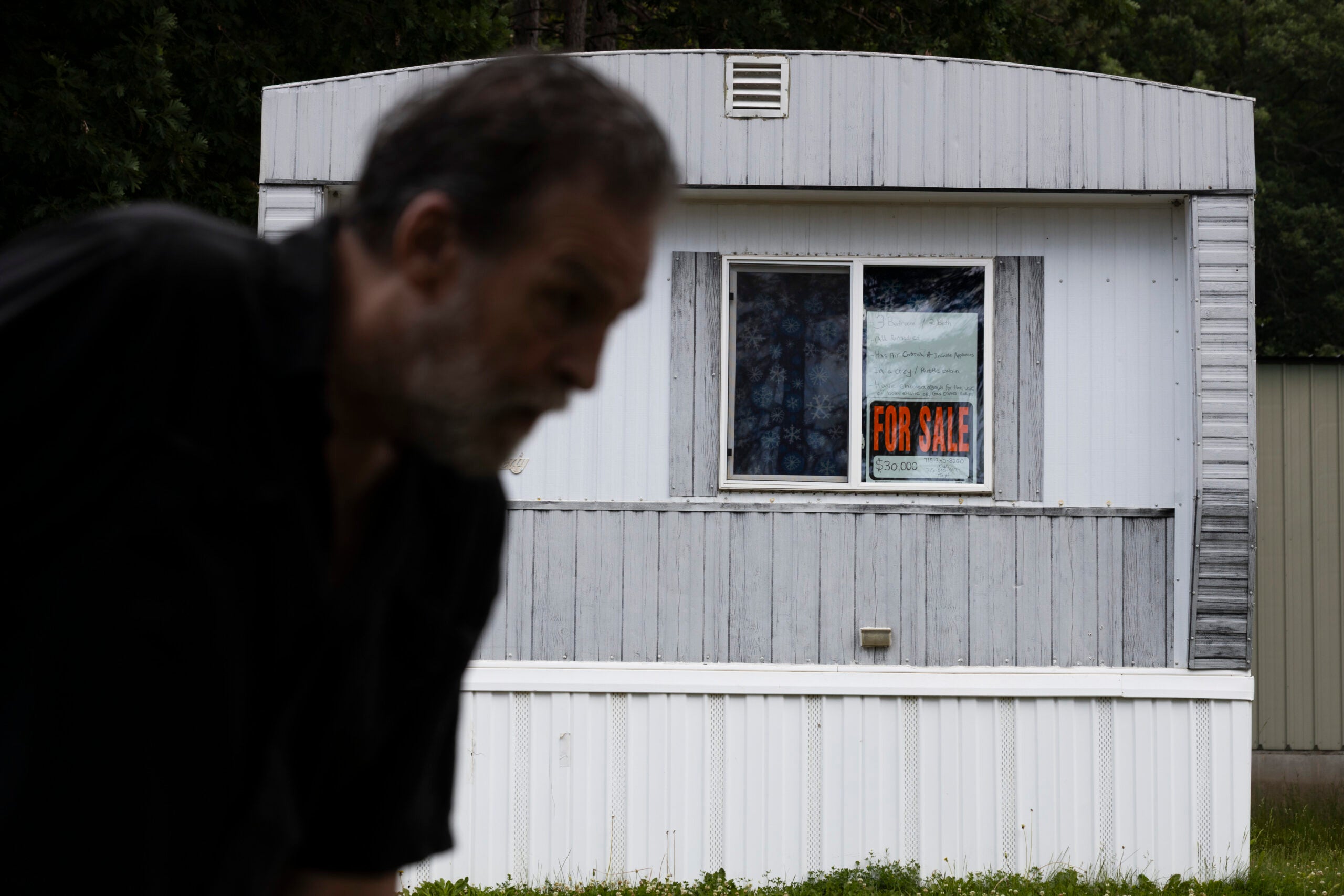 A blurred man in the foreground with a white mobile home in the background displaying a For Sale sign in its window.