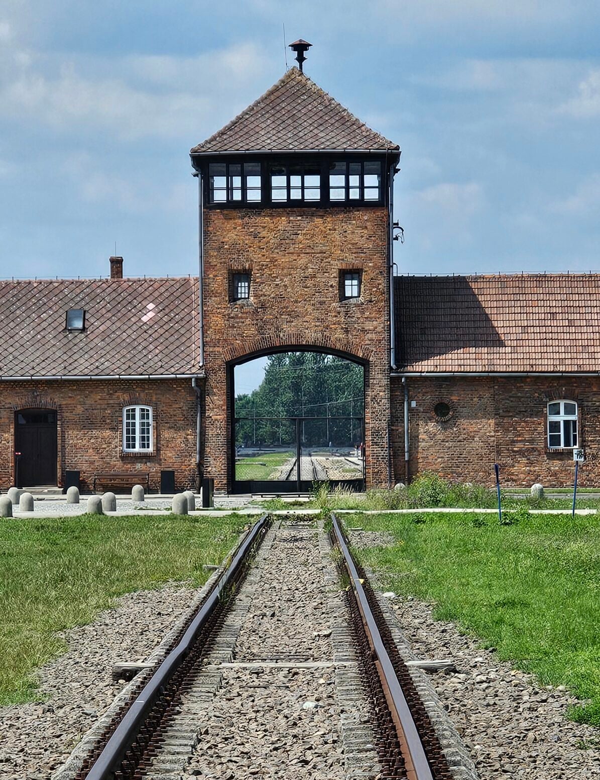 Railway tracks leading to the main entrance gate and guard tower of Auschwitz concentration camp, with brick buildings and grass on either side.
