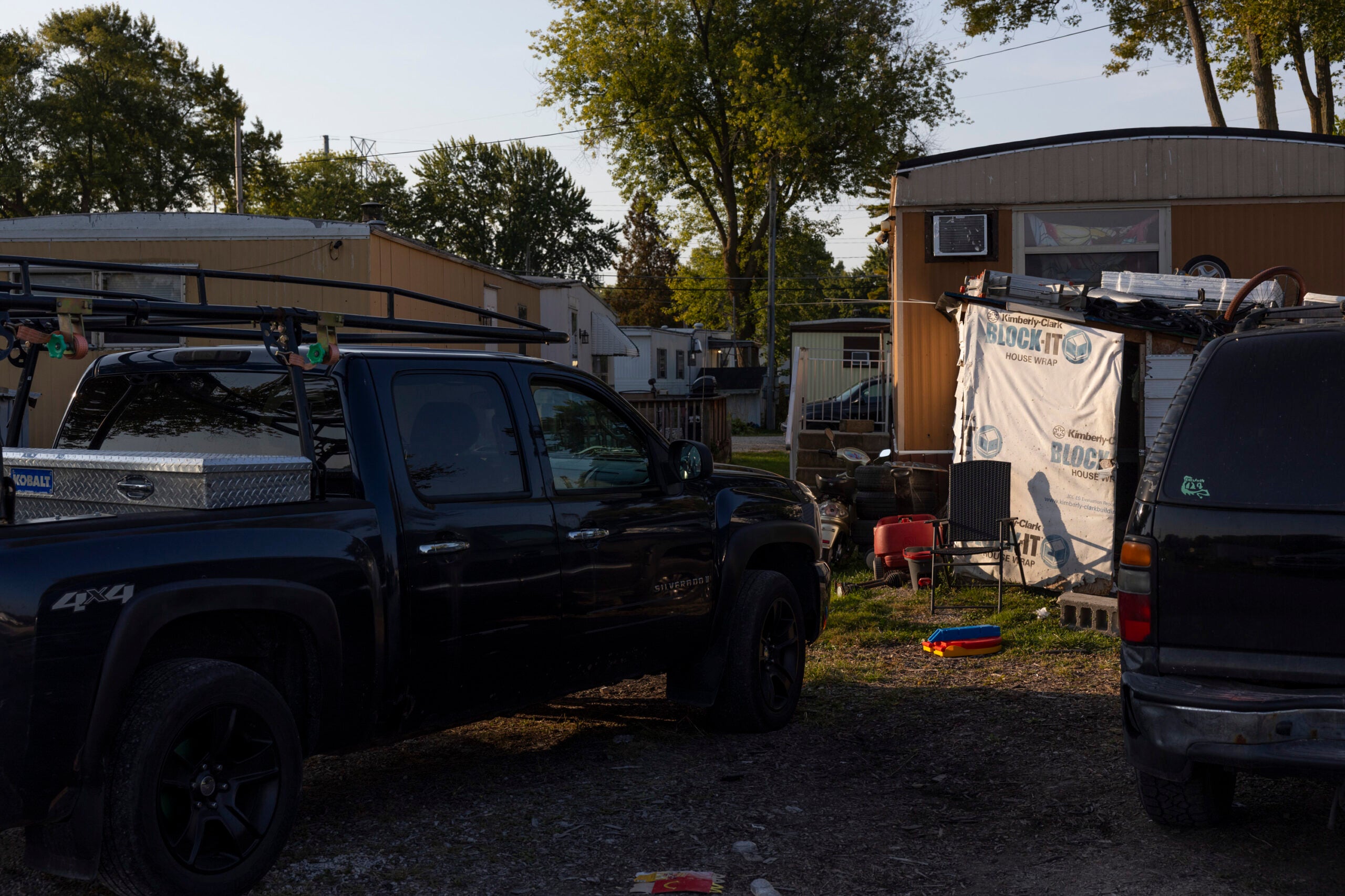 Two parked vehicles in front of a trailer home with a makeshift tarp covering, outdoor chairs, and scattered toys on the ground. Trees and additional trailers are in the background.