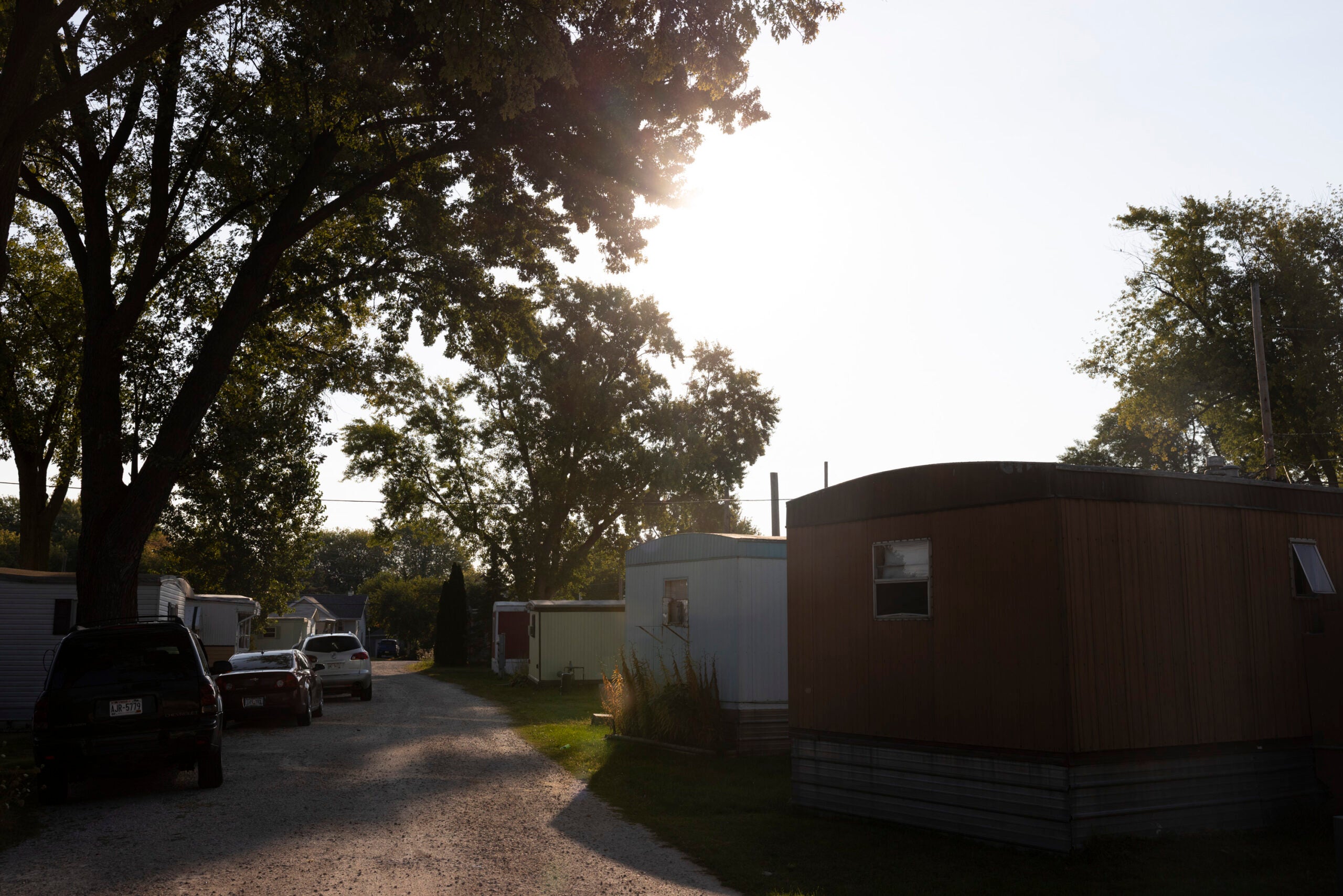 A trailer park with several mobile homes, cars parked on a gravel road, and sunlight shining through tall trees.