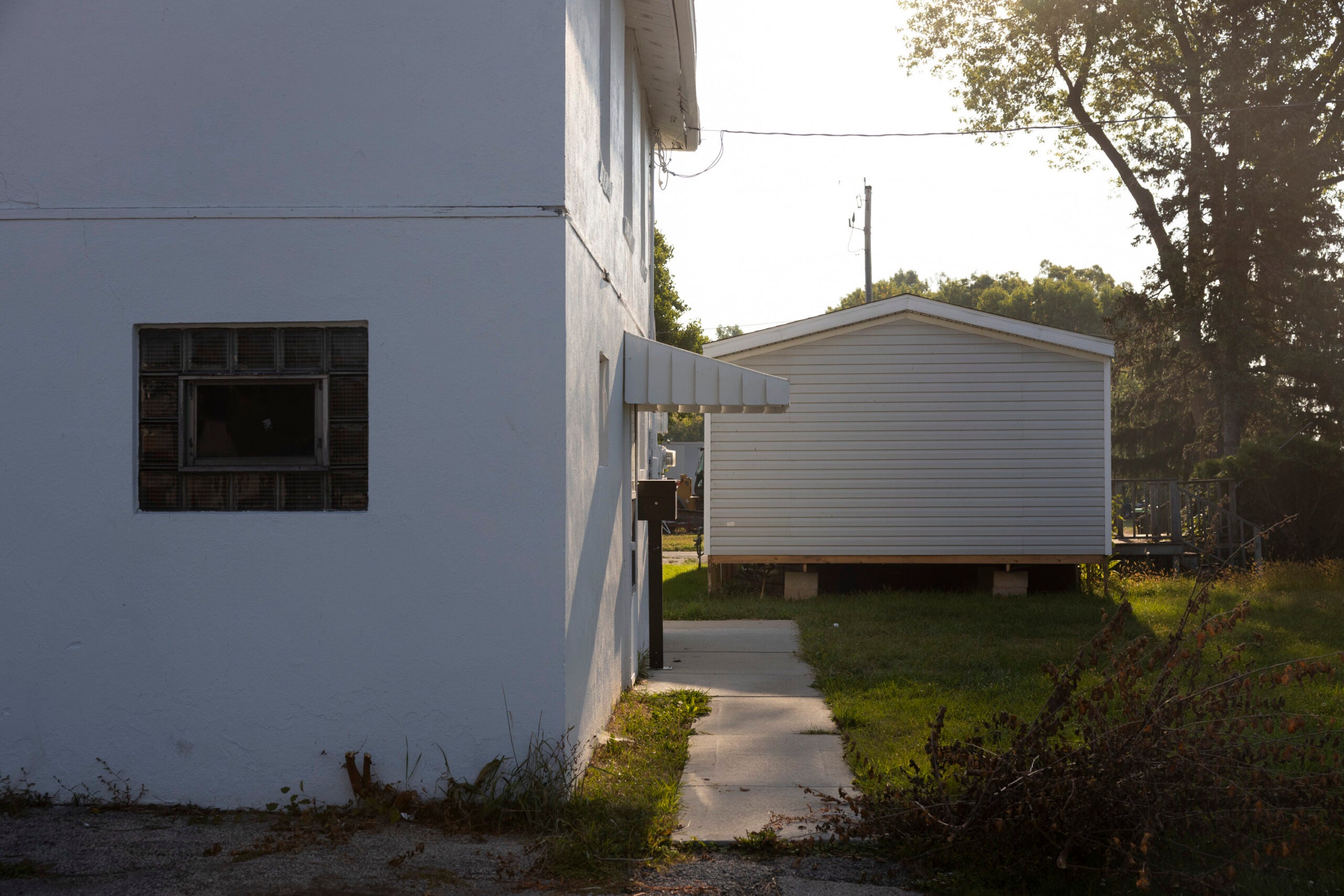 A white building with a small window and awning stands beside a white shed on a grassy lawn, separated by a concrete walkway under afternoon sunlight.