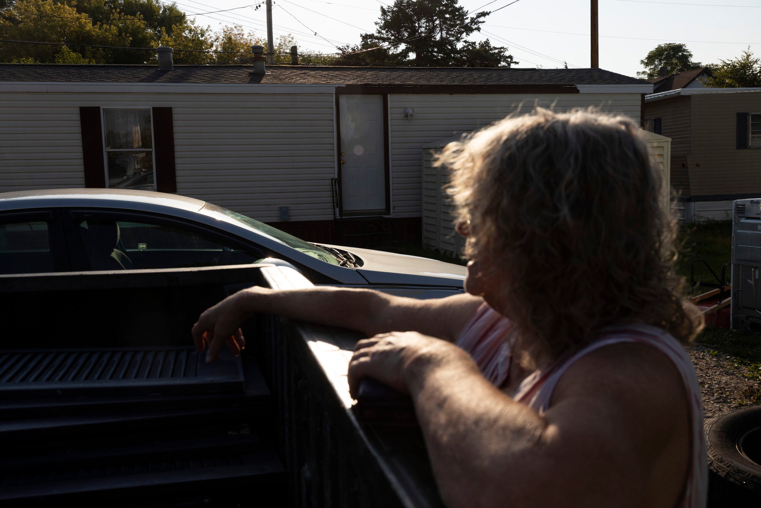 A person with wavy gray hair leans on the side of a truck bed near a white car, with a mobile home in the background, lit by late afternoon sun.