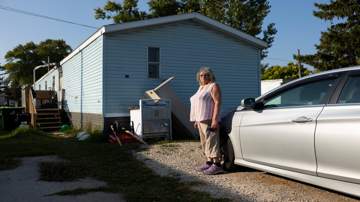 A woman stands next to a silver car parked beside a light blue mobile home with appliances and other items near the house.
