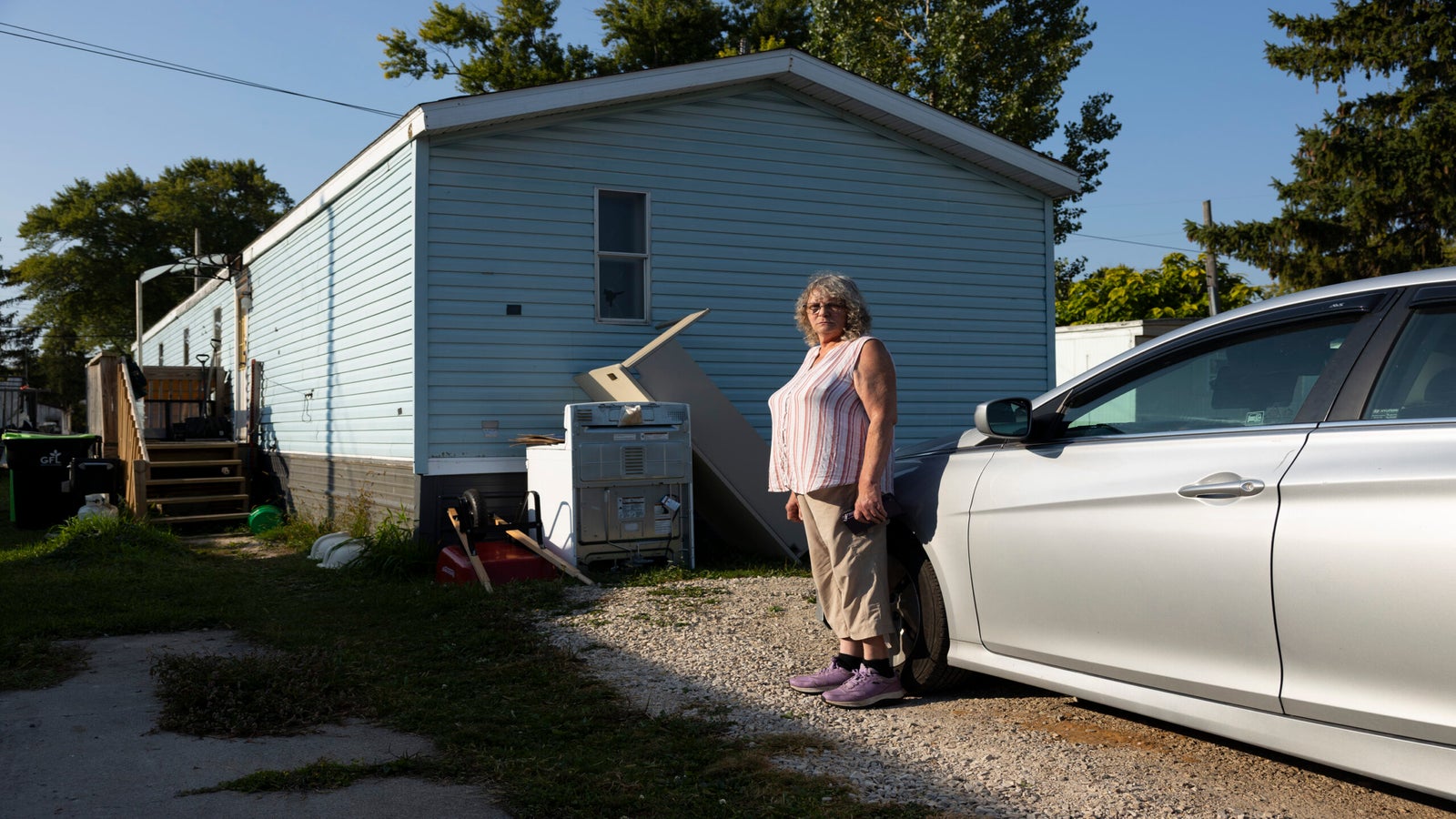 A woman stands next to a silver car parked beside a light blue mobile home with appliances and other items near the house.