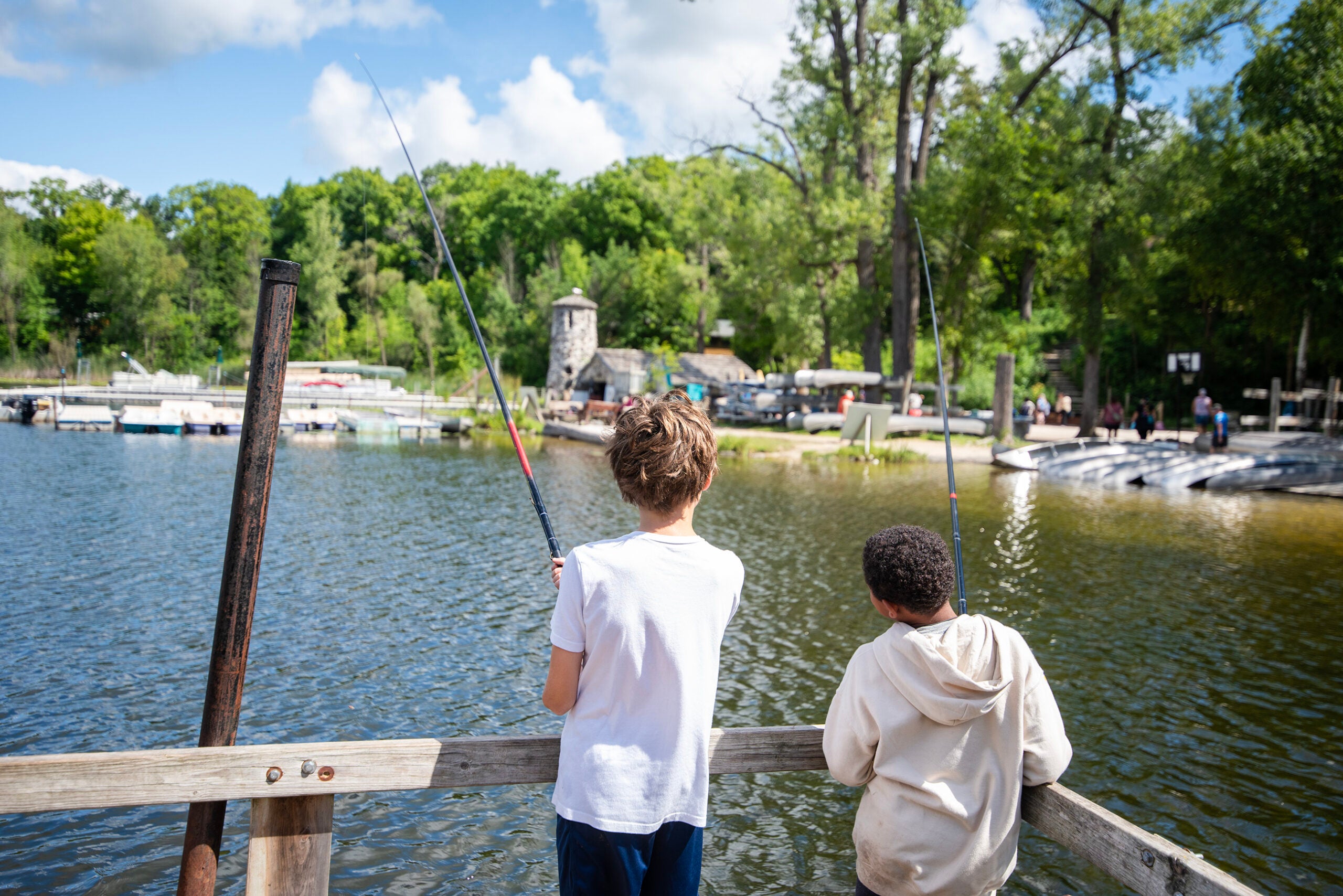 Two children stand on a wooden dock fishing in a lake, surrounded by trees and park buildings on a sunny day.