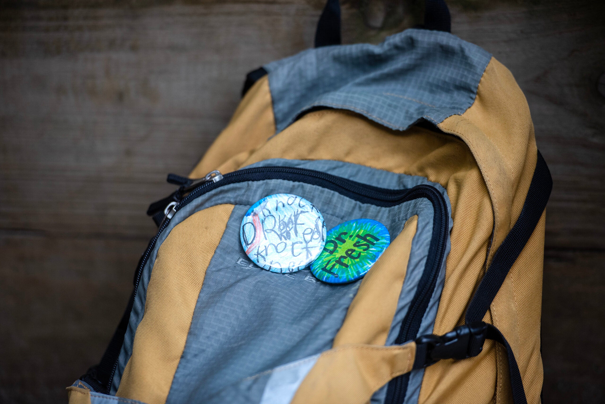 A yellow and gray backpack with two round, hand-decorated buttons attached to a zippered pocket, one featuring abstract text and the other reading for the trees.