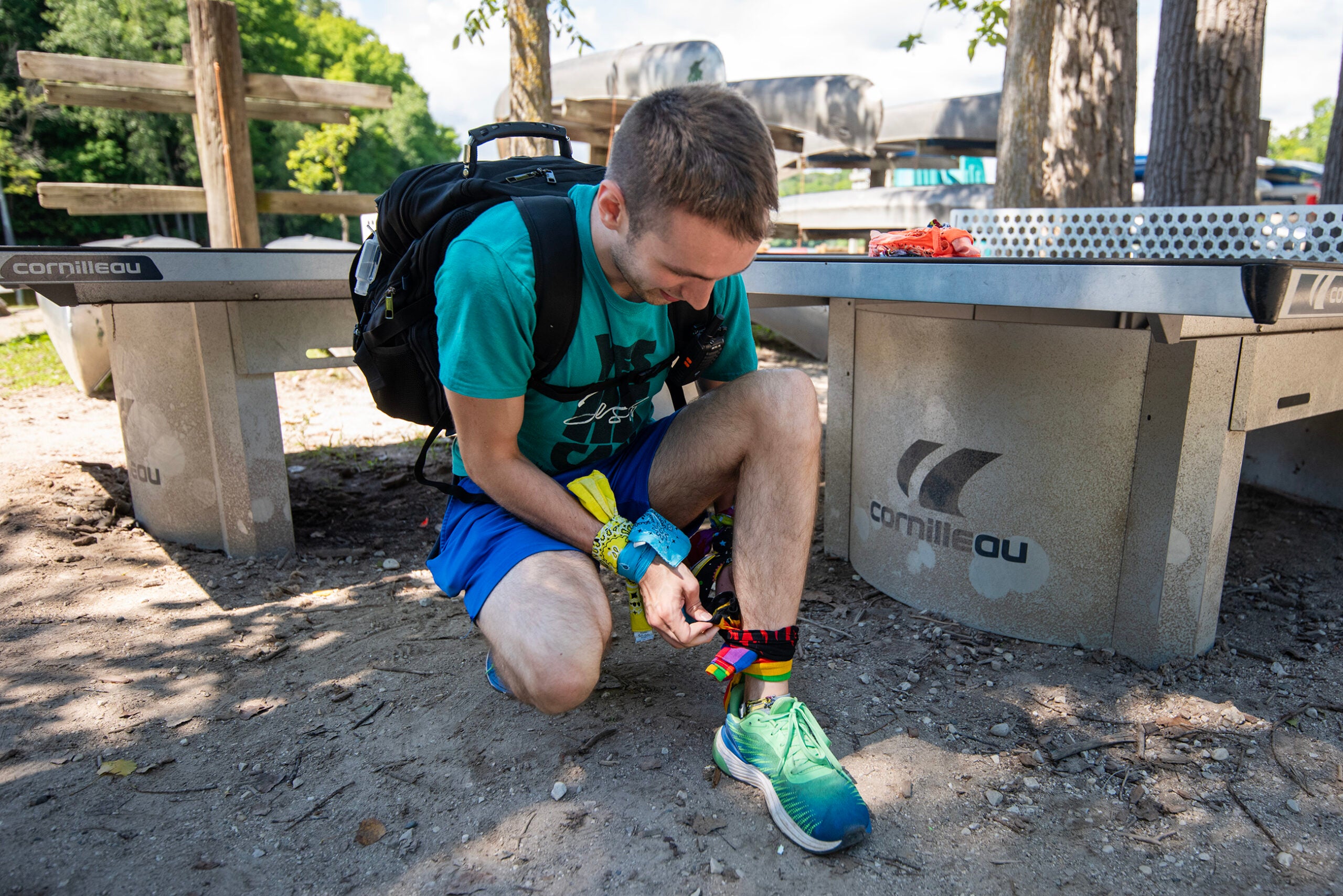 A person kneels by an outdoor ping pong table, wearing a backpack and athletic clothing, tying a colorful band around their ankle.