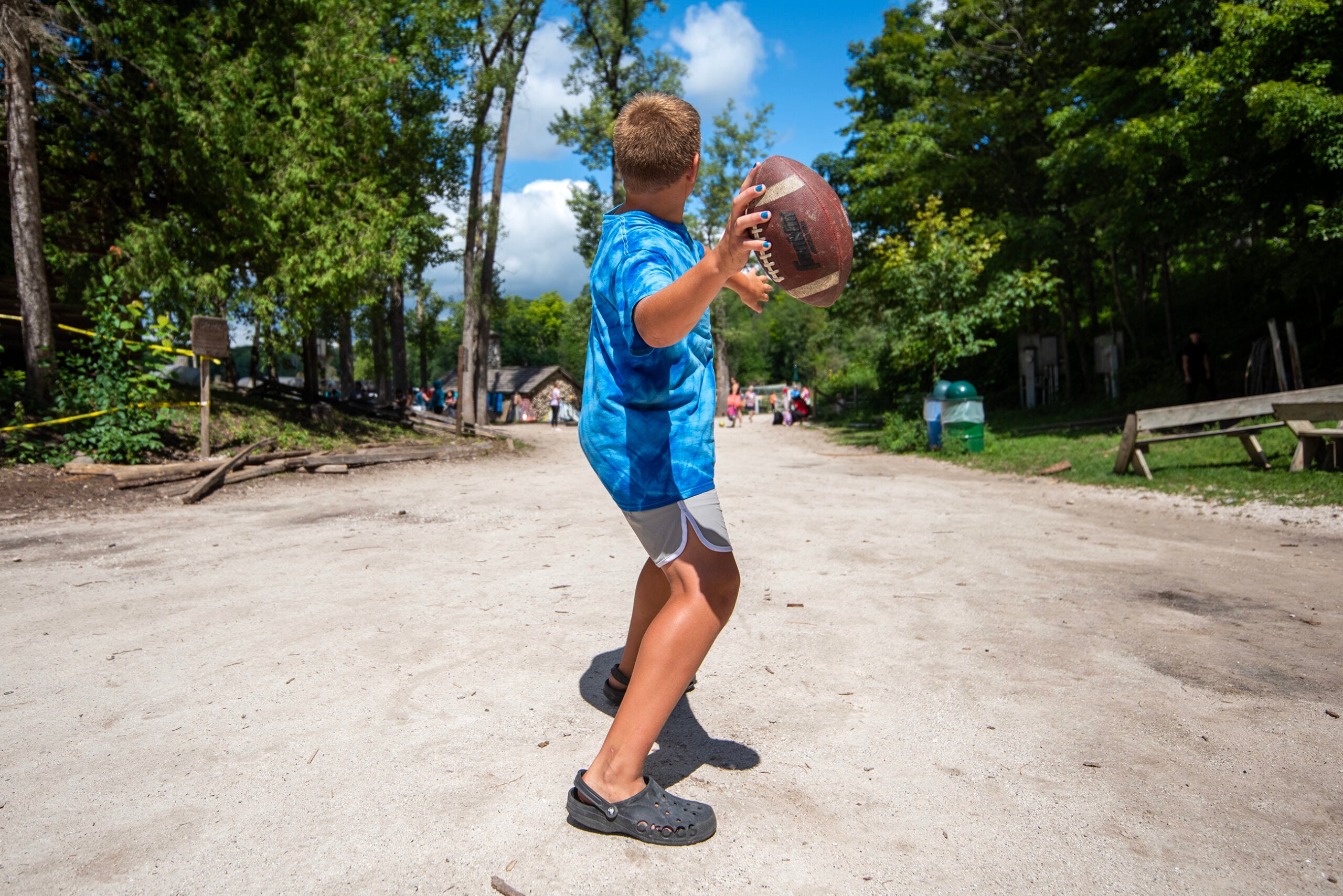 A boy in a blue tie-dye shirt stands on a dirt path, holding a football and preparing to throw it, with trees and people visible in the background.