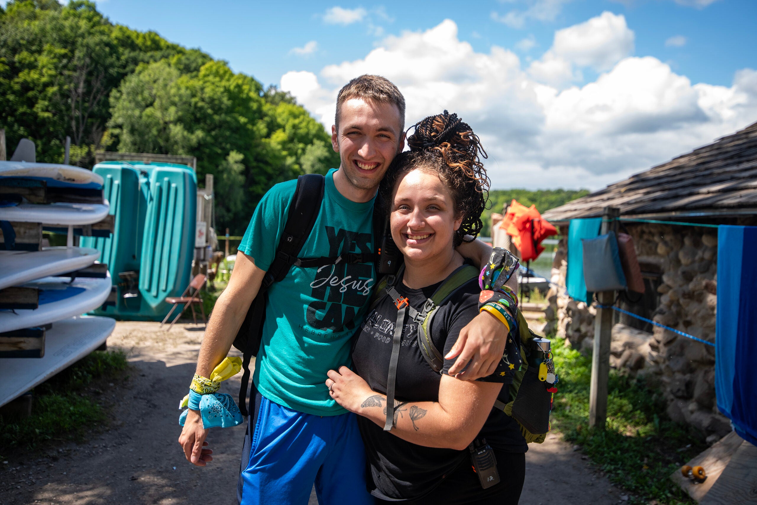 Two people stand close together smiling outdoors, with paddleboards, portable toilets, and trees visible in the background under a partly cloudy sky.